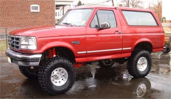 1994 Ford Bronco - Driver-side front three-quarter view of a red 1992-1996 Ford Bronco full-size SUV with a suspension lift, riding on chrome eight-hole wheel…