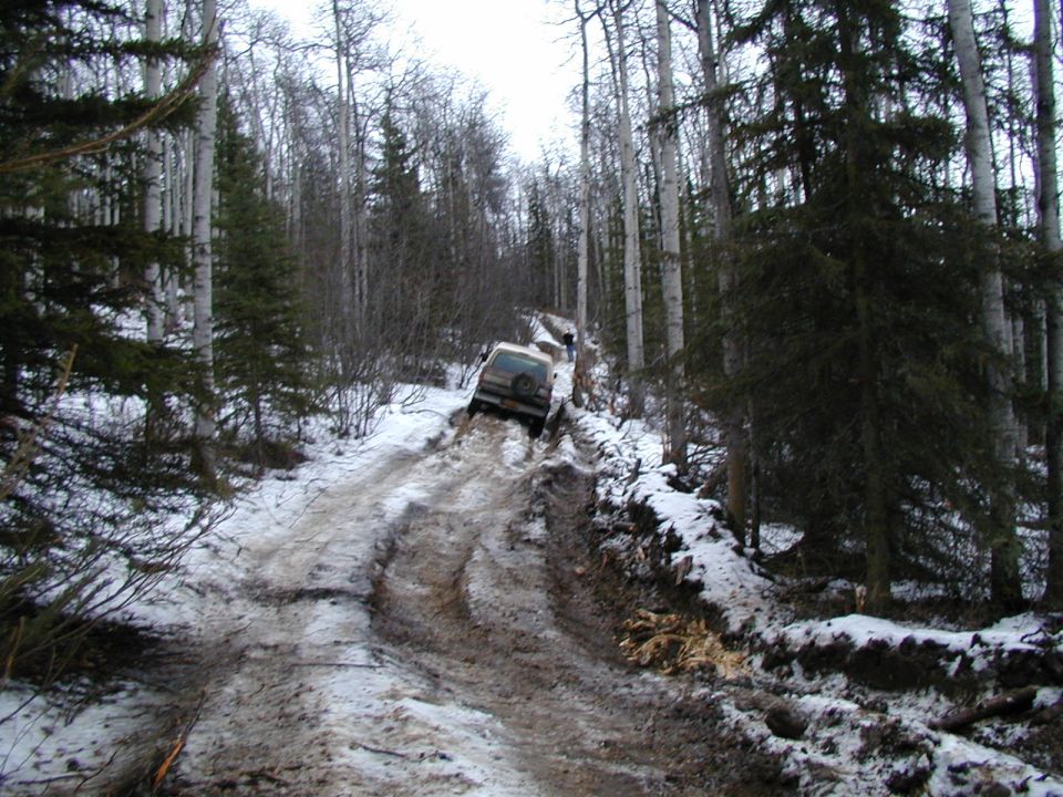 Light-colored SUV with rear spare tire on a muddy snowy forest trail, rear view