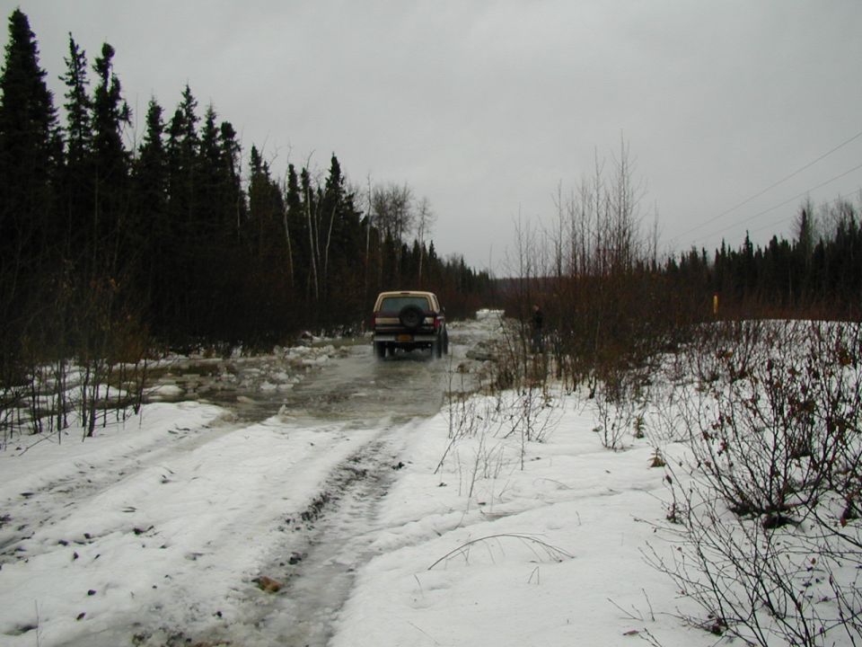 Two-tone SUV with rear spare tire crossing icy water on a snowy trail, viewed from behind