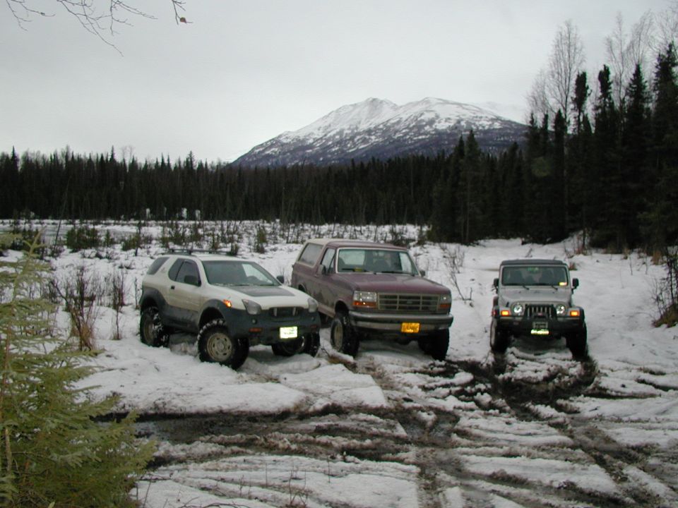 Silver Isuzu VehiCROSS, maroon Ford Bronco and silver Jeep Wrangler on mud tires in a snowy mountain clearing