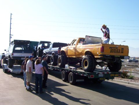 2000 Ford F-450 - Several lifted trucks are transported on a trailer, with a man standing in the bed of the yellow Ford.