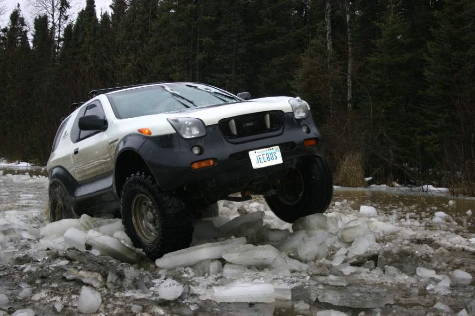 Lifted white/gray Isuzu Vehicross on chunky off-road tires perched on broken ice, forest behind.