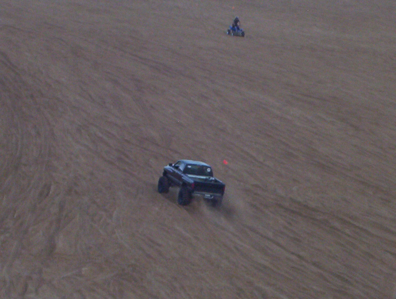 Lifted dark pickup on big tires climbing dune with ATV in background