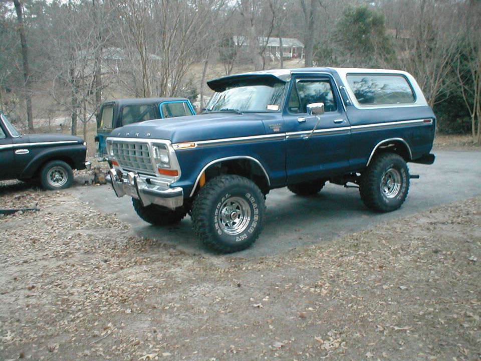 Lifted dark blue 1979 Ford Bronco with white top and big General Grabber tires on a gravel driveway
