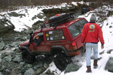 2003 Jeep TJ - A red Jeep Cherokee XJ drives through a challenging rocky and snowy landscape, with another vehicle visible in the background.
