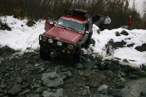 2003 Jeep TJ - A red Jeep Cherokee XJ navigates a rocky, snow-covered trail, with another vehicle visible in the background.