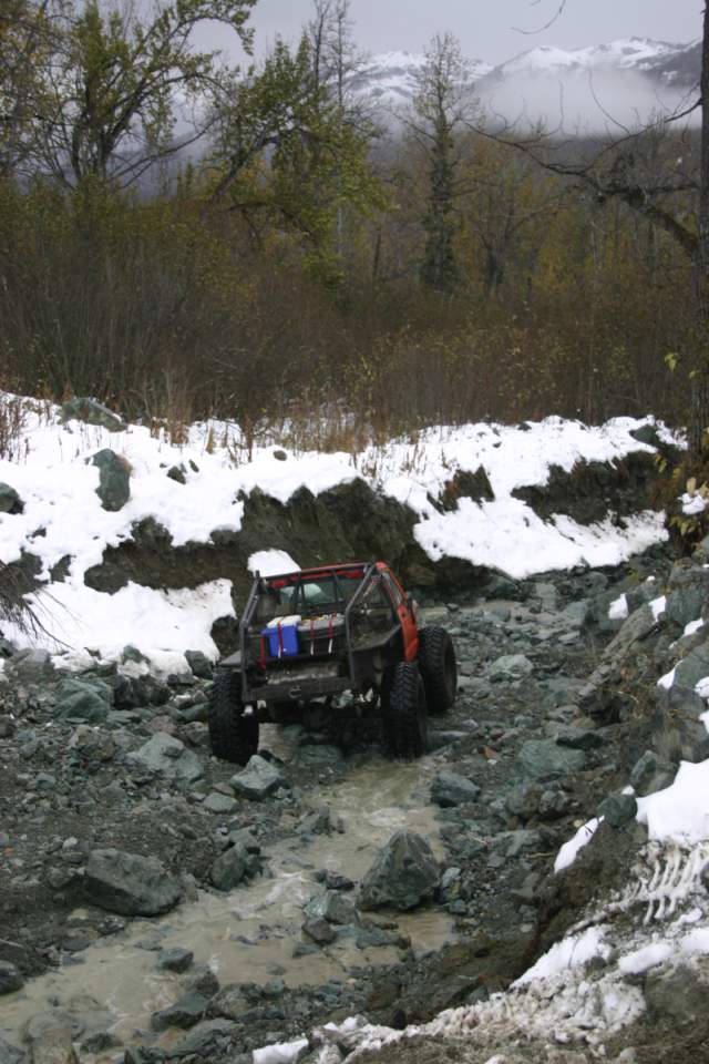 Orange off-road vehicle with a roll cage driving through a rocky, wet terrain with snow patches