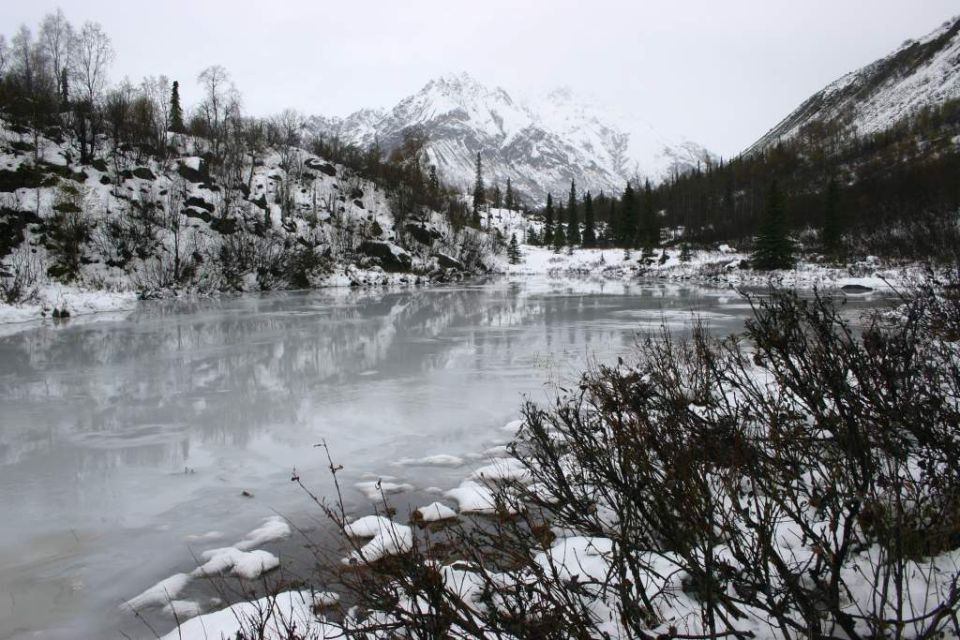 Frozen lake with reflections, surrounded by snow-covered hills and mountains under an overcast sky