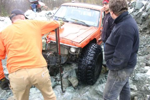 2003 Jeep TJ - An orange off-road vehicle is stuck in a rocky stream bed, with several people attending to it. The windshield displays 'ALASKA OFFROAD'.