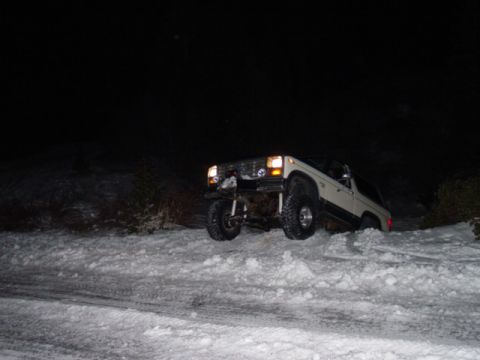 1986 Ford Bronco - A white SUV navigates a snowy hill in the dark, with its headlights illuminating the path.