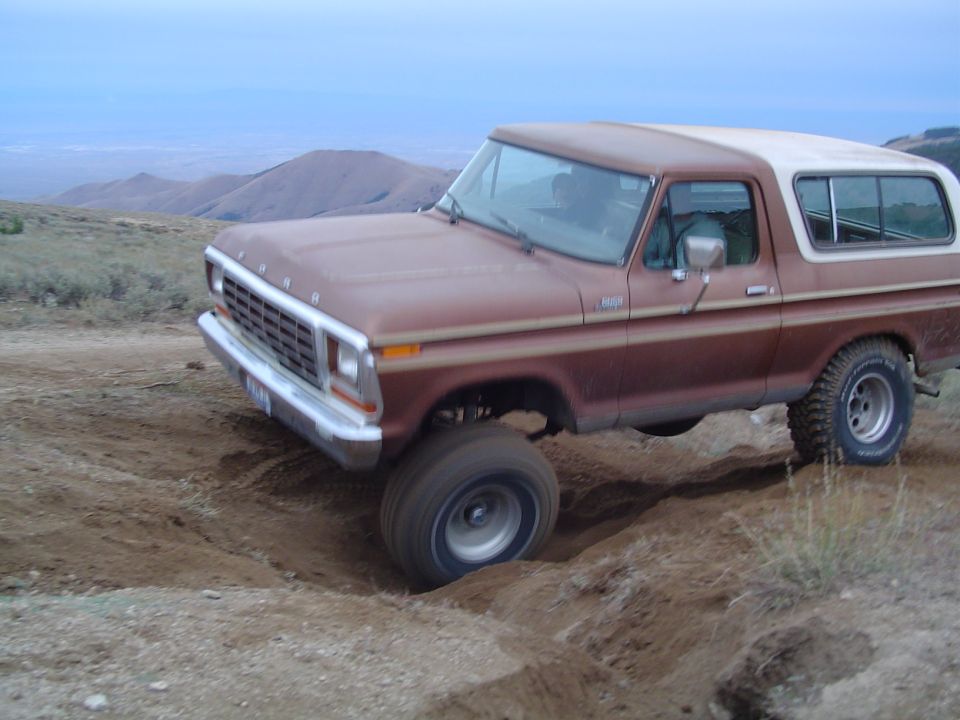 Brown 1978 Ford Bronco with a white top driving off-road, front-left wheel in a ditch