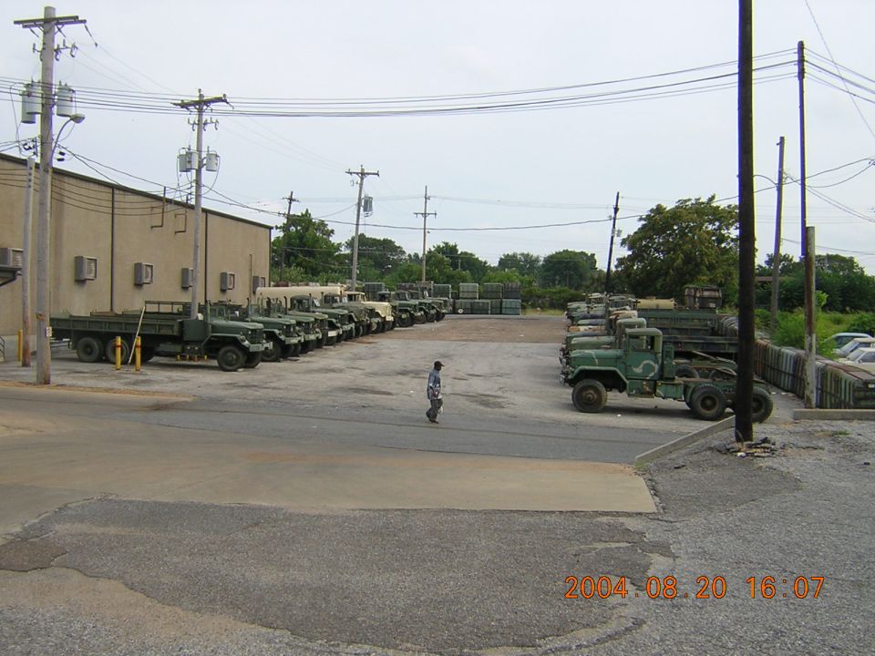 Rows of olive drab military cargo trucks parked in a fenced surplus lot, 2004