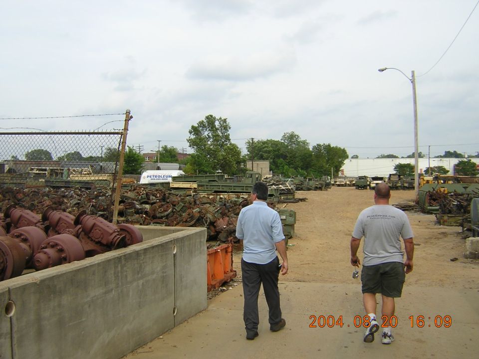 Two men walk through a junkyard filled with vehicle parts and military-style trucks