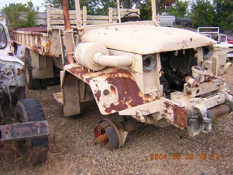 1984 Chevrolet Blazer - A weathered tan military truck sits in a junkyard, its front end heavily damaged and engine exposed, with another vehicle partially visible…