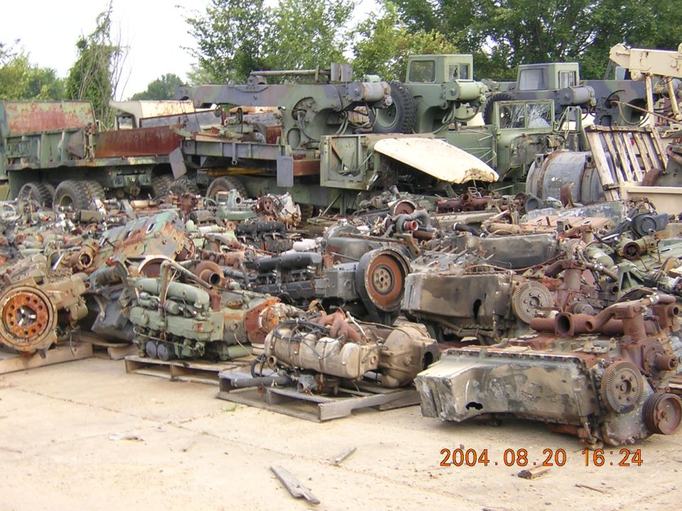 Pile of various engines and truck parts, some on pallets, with military trucks in the background