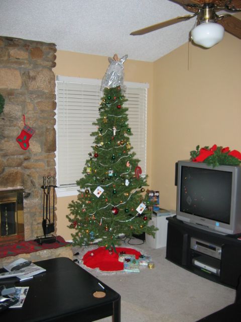 1985 Ford LTD Crown Victoria - A decorated Christmas tree stands in a living room next to a stone fireplace and a television.
