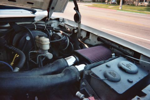 1990 Ford Bronco - engine bay with a red cone air filter, master-cylinder reservoir, and battery, photographed with the hood up and a street in the background.