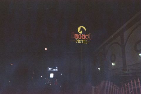 1990 Ford Bronco - lit Bronco Motel sign glows in the dark night sky above a street sign marked 55th St, with a building railing at right.