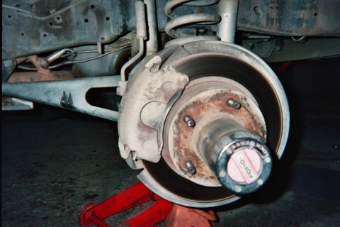 1990 Ford Bronco - vehicle's front disc brake rotor, caliper, and rusty hub are exposed during service, with a Ford-stamped grease cap and a red jack below.