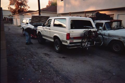 1990 Ford Bronco - Two white Ford Broncos and a weathered older pickup sit in an alley at dusk while a person crouches working at a front wheel.