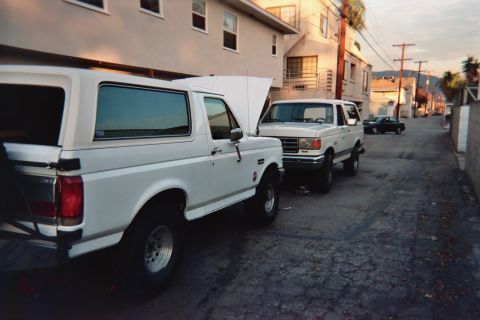 1990 Ford Bronco - Two white Ford Broncos, one with a hardtop, park nose-to-tail down a residential alley at dusk with utility poles and hills in the distance.
