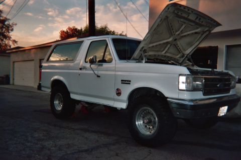 1990 Ford Bronco - white lifted Ford Bronco with its hood raised sits in an alley at sunset, oversized tires and chrome grille catching the warm light.