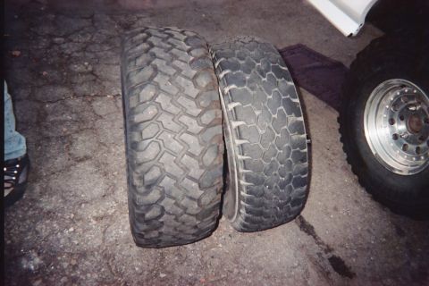 1990 Ford Bronco - Two worn off-road tires with different tread patterns lean side by side on dark pavement, a mounted wheel and white vehicle body at right.