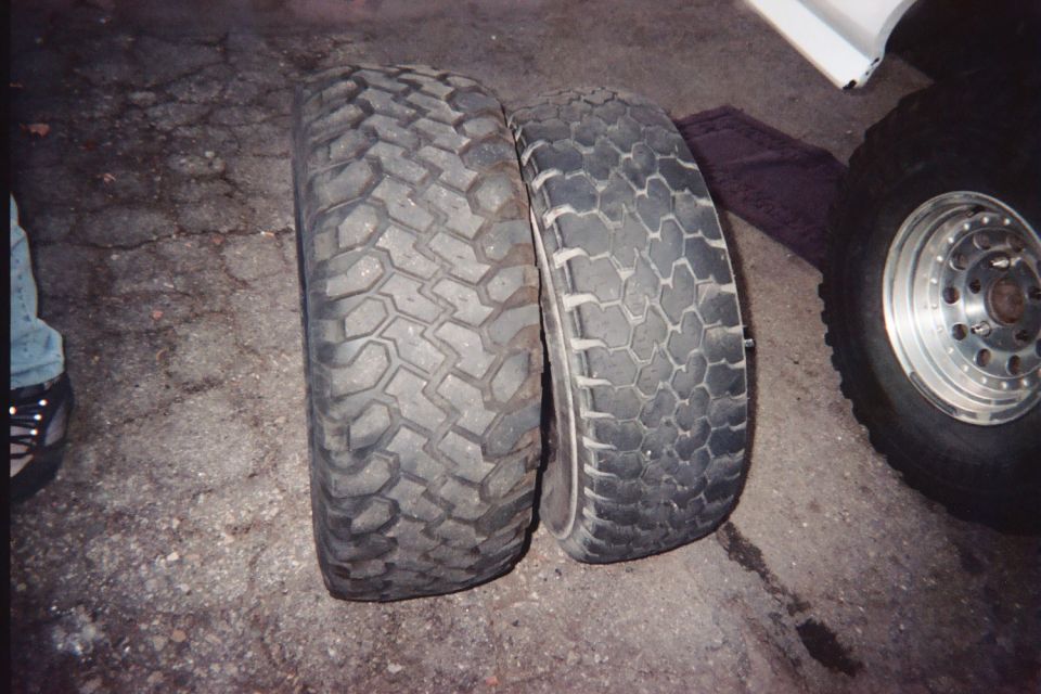 Two off-road tires of different tread patterns stood side by side on pavement beside a wheel