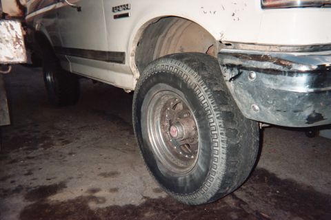 1990 Ford Bronco - tire on a chrome-look wheel leans against the front fender of a white Ford truck inside a dim garage, exposing the bare hub behind.