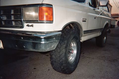 1990 Ford Bronco - front corner of a white Ford 4x4 truck, with amber turn-signal lens, chrome bumper, 4x4 badge, and a large off-road tire on a polished whee…