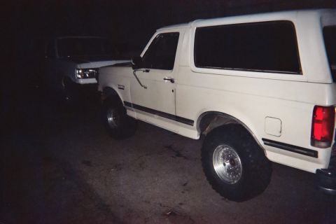 1990 Ford Bronco - Two white Ford Broncos parked in the dark, the foreground one lifted on oversized tires with its red taillight catching the flash.
