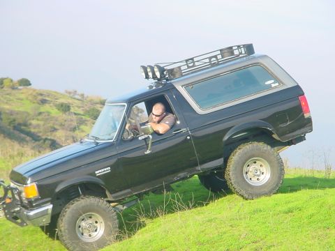 1988 Ford Bronco - A black Ford Bronco with off-road modifications navigates a steep, grassy incline under a clear sky.