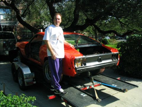 1969 Ford Mustang - A man stands beside an orange 1969 Ford Mustang being transported on a trailer.