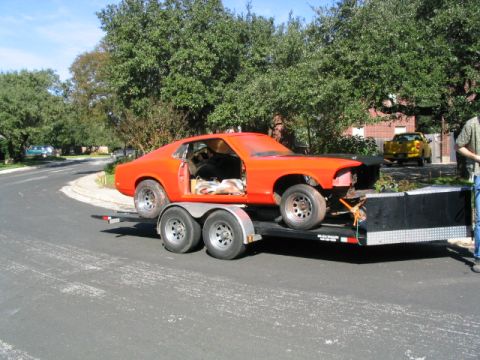 1969 Ford Mustang - An orange 1969 Ford Mustang coupe is transported on a trailer, with a yellow pickup truck visible in the background.