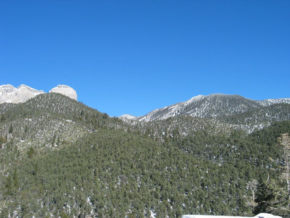 Snow-dusted evergreen trees cover mountains under a clear blue sky. Watermark reads SUPERMOTORS.NET