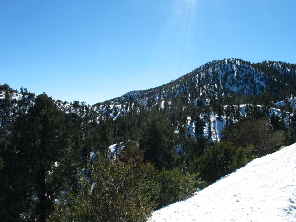 Snowy mountain landscape with pine trees under a clear blue sky. Watermark reads SUPERMOTORS.NET