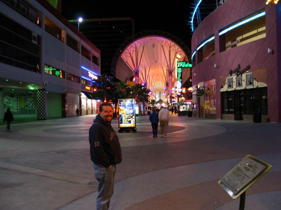 Man in a dark hoodie and jeans stands in a plaza at night with neon lights and buildings in the background