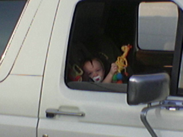 Baby with pacifier and giraffe toy in side window of white vehicle