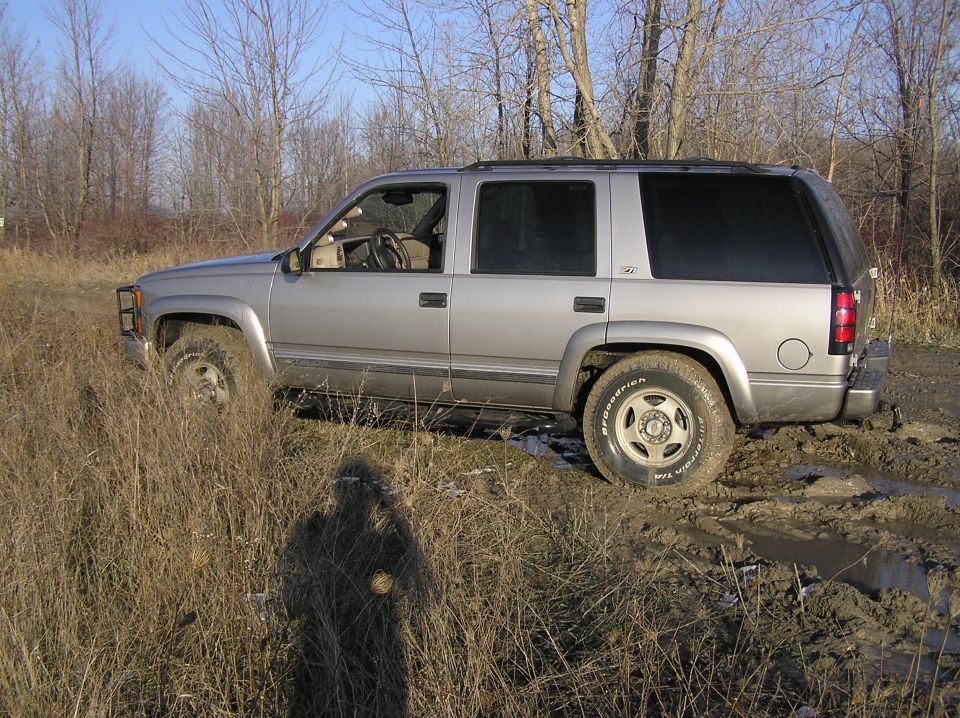 Silver SUV stuck in mud, side view, with tall dry grass in foreground and bare trees in background