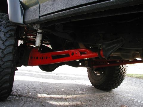 1994 Ford Bronco - Low ground-level view under the front of a lifted truck showing red powder-coated lift-kit radius arms with drilled lightening holes, a whi…
