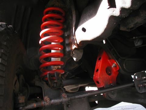 1994 Ford Bronco - Close-up under a 4x4 front suspension showing a red aftermarket lift coil spring seated over the shock, with a red multi-hole drop bracket…