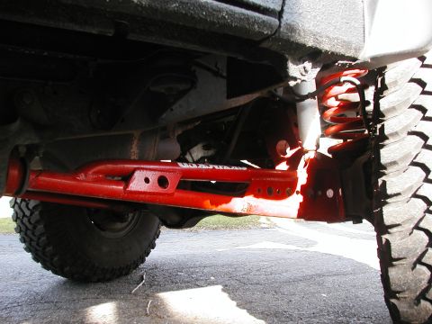 1994 Ford Bronco - Under-vehicle close-up of a red powder-coated tubular radius arm with drilled holes and white lettering, bolted to the front axle bracket o…