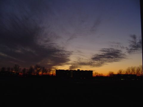 1990 Ford Bronco - A dramatic sunset paints the sky with hues of purple, orange, and yellow over a silhouetted landscape of buildings and trees.