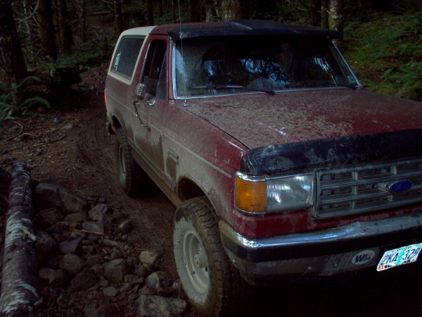 Red Ford Bronco with a white top driving on a muddy, rocky forest trail. License plate visible