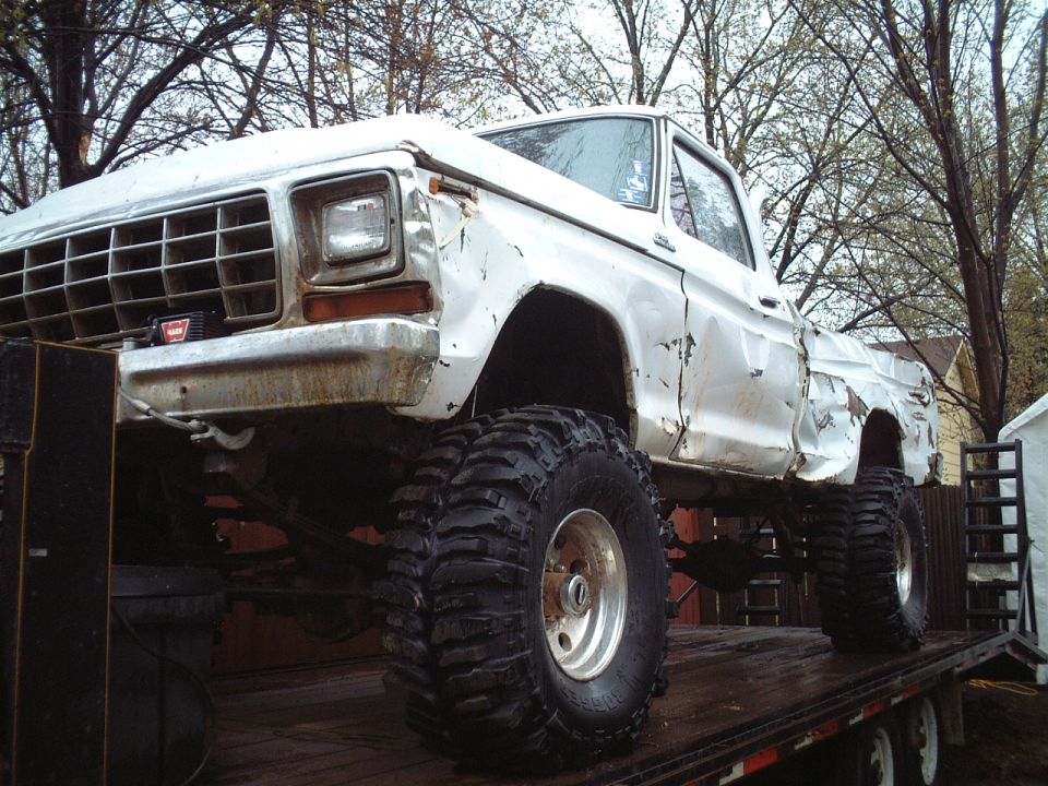 White 1979 Ford F-150 pickup truck with large off-road tires, dented body panels, on a flatbed trailer