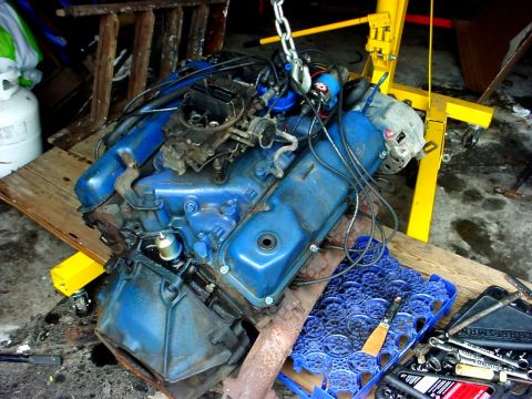 1973 Ford Ranchero - A blue V8 engine is hoisted by a yellow engine crane, resting on a wooden platform in a garage.