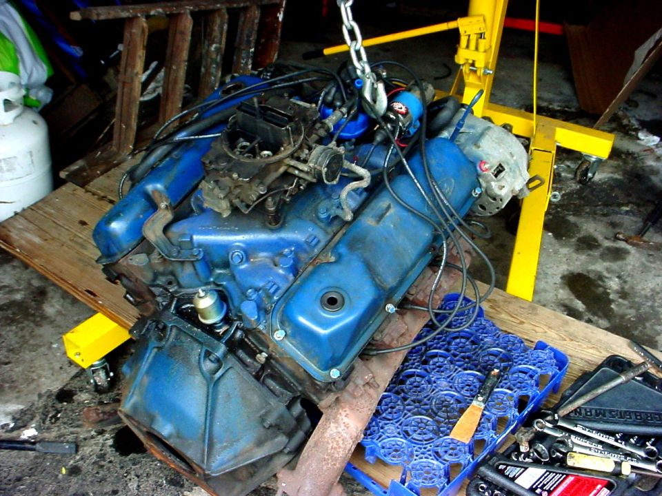 Blue V8 engine on a wooden platform, suspended by a yellow engine hoist