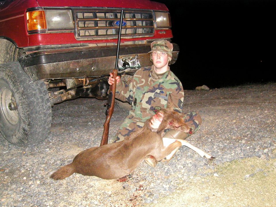 Red truck front end with a person holding a deer and rifle