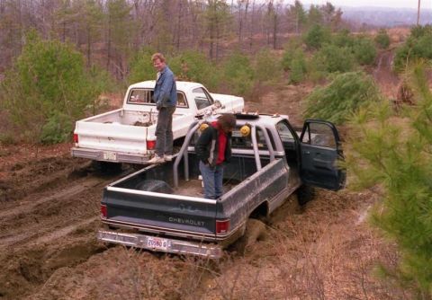 1991 Ford Bronco - Two Chevrolet pickup trucks are stuck in the mud off-road, with two men standing in the beds.