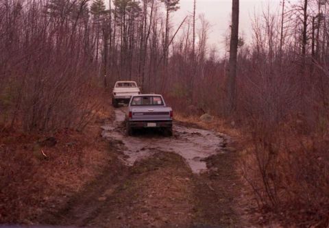 1991 Ford Bronco - Two pickup trucks navigate a muddy path through a wooded area.